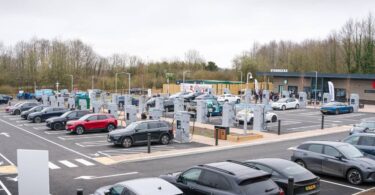 Large EV charging hub with multiple ultra-rapid chargers and electric vehicles parked at bays, located beside a retail site in the UK.