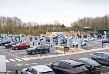 Large EV charging hub with multiple ultra-rapid chargers and electric vehicles parked at bays, located beside a retail site in the UK.