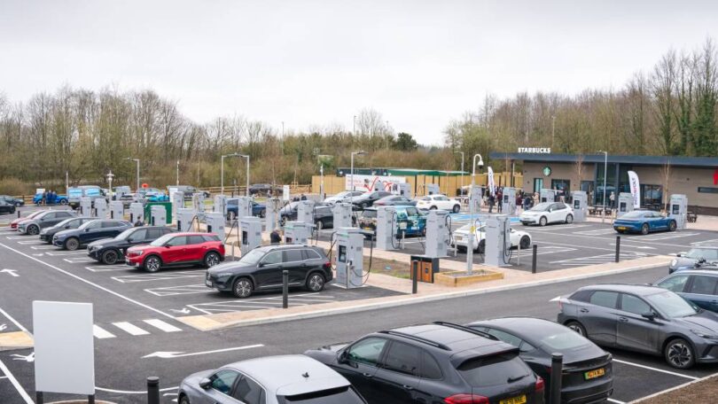 Large EV charging hub with multiple ultra-rapid chargers and electric vehicles parked at bays, located beside a retail site in the UK.