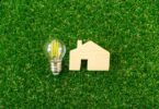 Wooden house model and LED lightbulb on green grass representing home energy efficiency and lower energy bills.