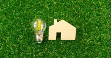 Wooden house model and LED lightbulb on green grass representing home energy efficiency and lower energy bills.