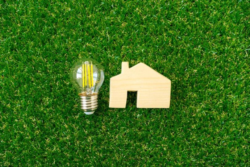 Wooden house model and LED lightbulb on green grass representing home energy efficiency and lower energy bills.