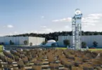 Rows of tilted mirror panels face a tall steel lattice tower with a white dish on top, beside large gray industrial buildings
