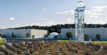 Rows of tilted mirror panels face a tall steel lattice tower with a white dish on top, beside large gray industrial buildings