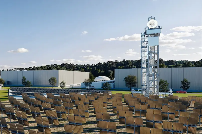 Rows of tilted mirror panels face a tall steel lattice tower with a white dish on top, beside large gray industrial buildings