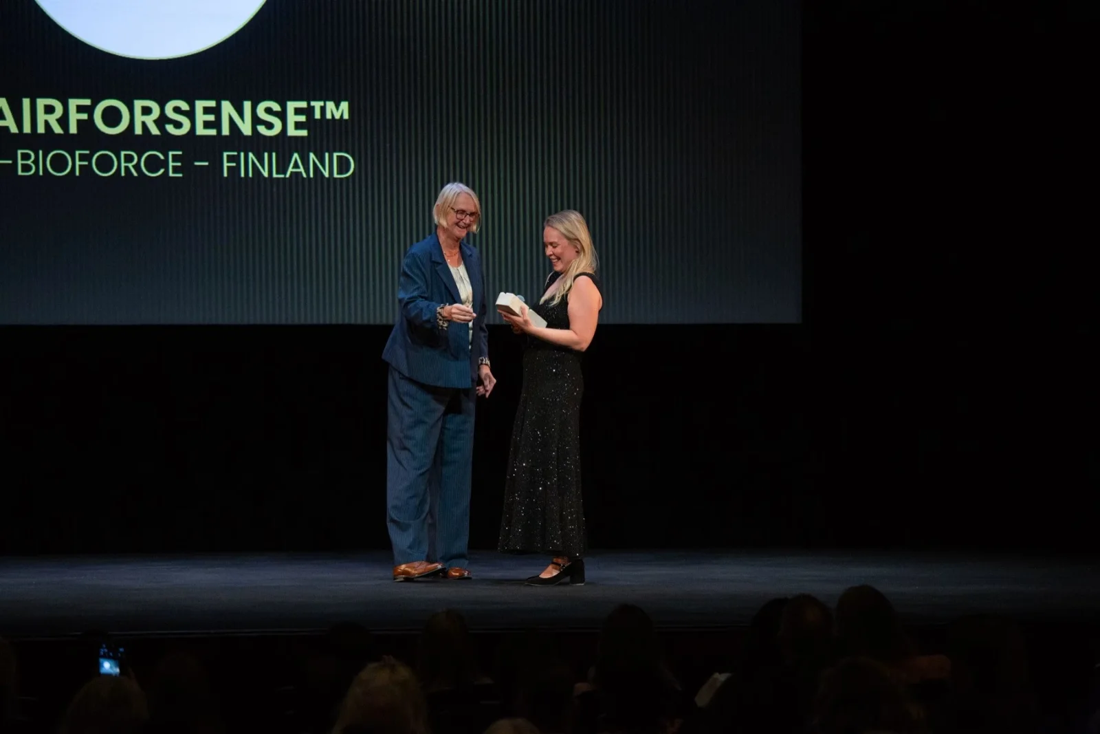 Two women on a dark stage beneath a screen reading AIRFORSENSE, BIOFORCE – FINLAND, one holding a small white box