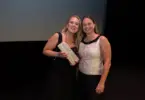 Two women on a dark stage beneath a screen reading AIRFORSENSE, BIOFORCE – FINLAND, one holding a small white box