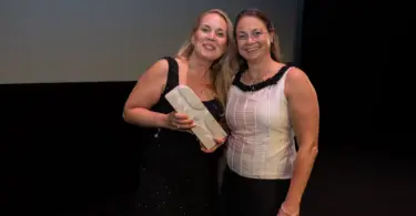 Two women on a dark stage beneath a screen reading AIRFORSENSE, BIOFORCE – FINLAND, one holding a small white box