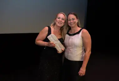 Two women on a dark stage beneath a screen reading AIRFORSENSE, BIOFORCE – FINLAND, one holding a small white box