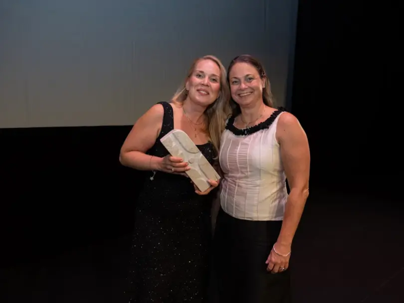 Two women on a dark stage beneath a screen reading AIRFORSENSE, BIOFORCE – FINLAND, one holding a small white box