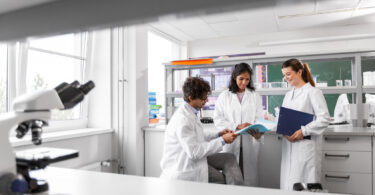Group of smiling scientists in a laboratory, one holding a folder, discussing a report together.