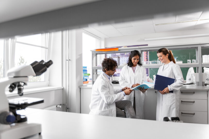 Group of smiling scientists in a laboratory, one holding a folder, discussing a report together.