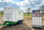 White HPU generator unit on a green trailer beside a 300 bar gas cylinder bundle on bare ground, with fields and trees behind