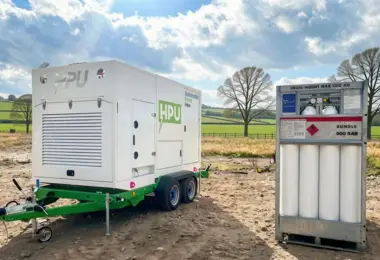 White HPU generator unit on a green trailer beside a 300 bar gas cylinder bundle on bare ground, with fields and trees behind