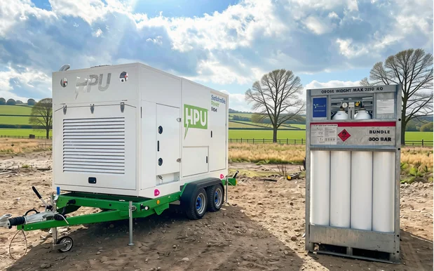 White HPU generator unit on a green trailer beside a 300 bar gas cylinder bundle on bare ground, with fields and trees behind