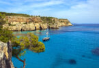 A ship sailing on clear turquoise water at Cala Macarella, Menorca, with a Mediterranean coastline in the background.