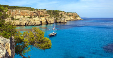 A ship sailing on clear turquoise water at Cala Macarella, Menorca, with a Mediterranean coastline in the background.