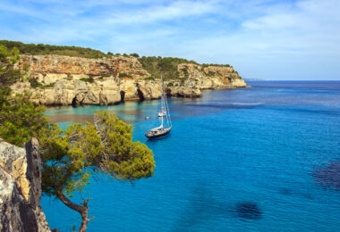 A ship sailing on clear turquoise water at Cala Macarella, Menorca, with a Mediterranean coastline in the background.