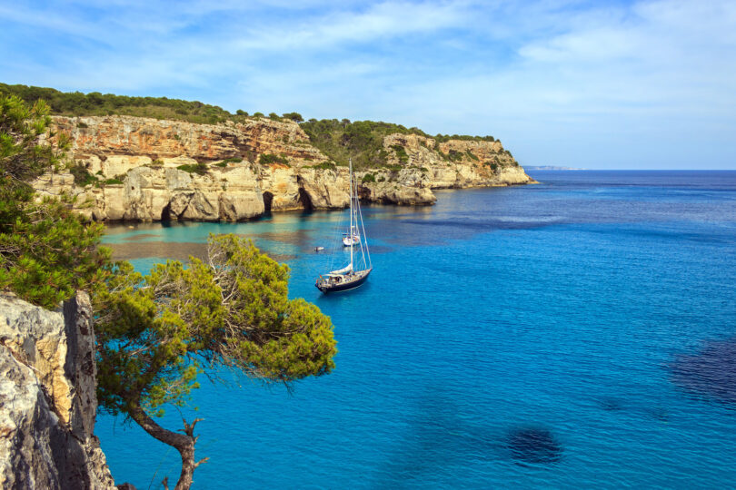 A ship sailing on clear turquoise water at Cala Macarella, Menorca, with a Mediterranean coastline in the background.
