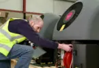 Man in hi-vis vest reaching into the open panel of a grey heat pump unit with a pink octopus logo on its fan, in a warehouse