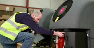 Man in hi-vis vest reaching into the open panel of a grey heat pump unit with a pink octopus logo on its fan, in a warehouse