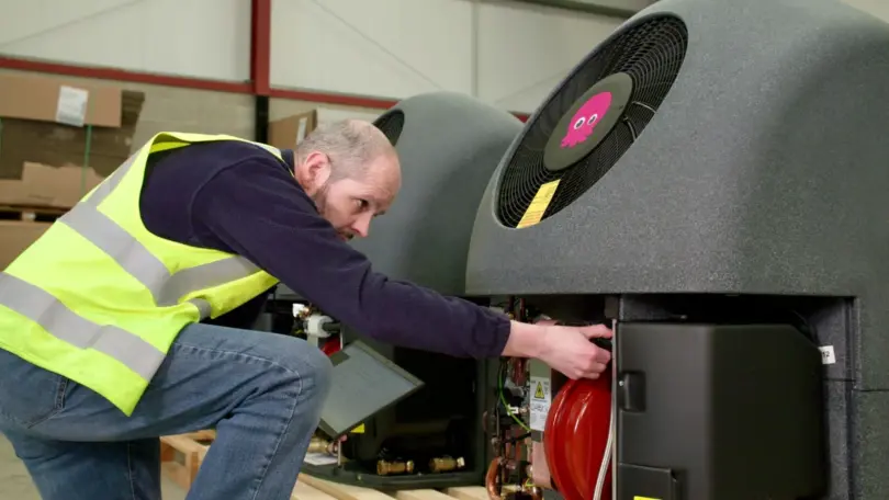 Man in hi-vis vest reaching into the open panel of a grey heat pump unit with a pink octopus logo on its fan, in a warehouse