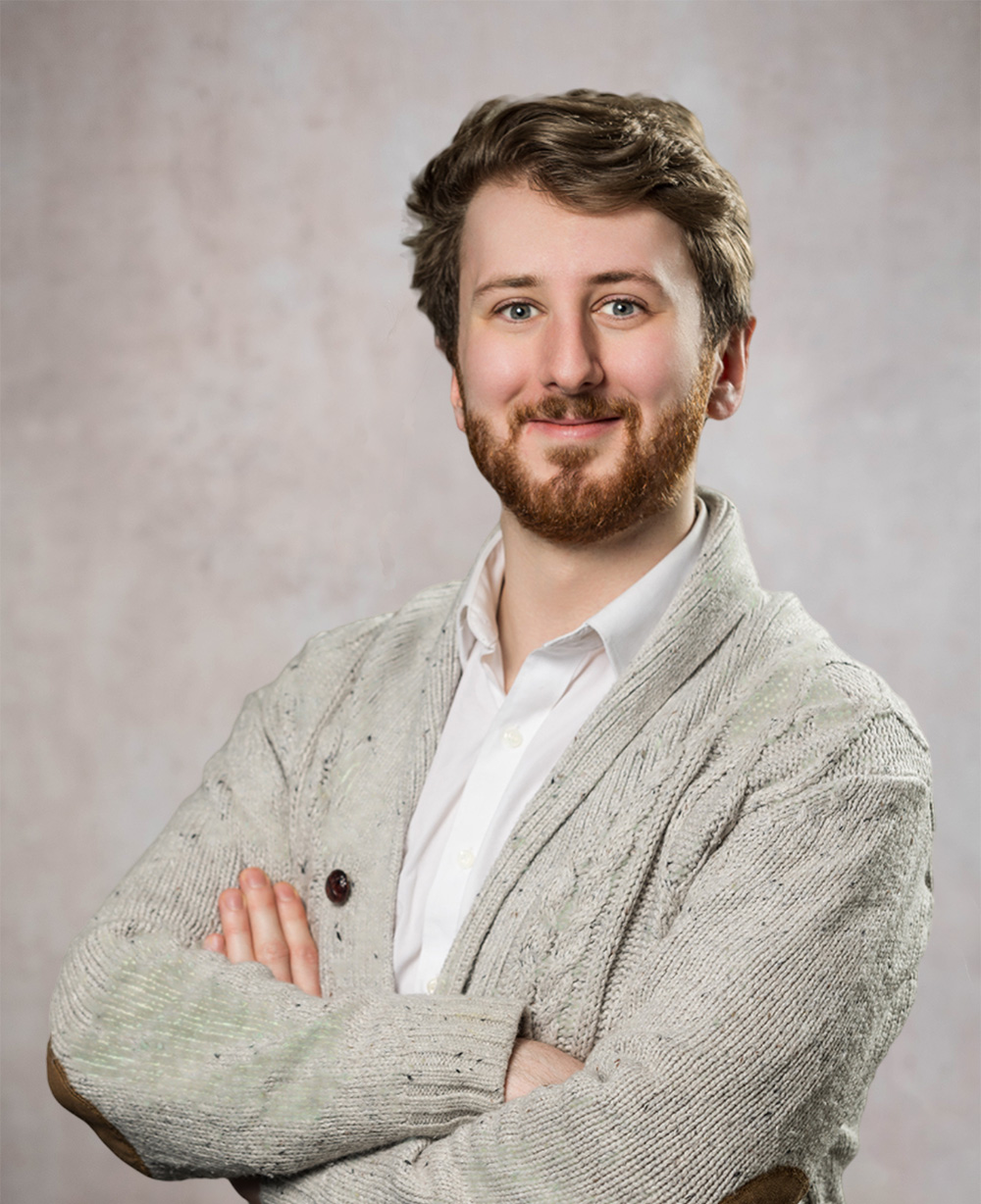 Smiling bearded man with swept-back brown hair, in a grey knit cardigan and white shirt, arms crossed against a mottled backd