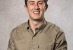 Smiling young man with short curly brown hair wearing a tan button-down shirt against a mottled gray backdrop