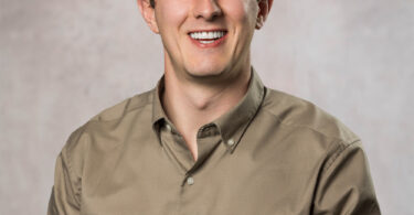Smiling young man with short curly brown hair wearing a tan button-down shirt against a mottled gray backdrop