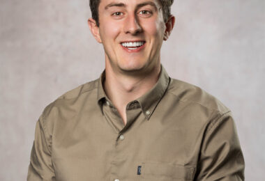 Smiling young man with short curly brown hair wearing a tan button-down shirt against a mottled gray backdrop