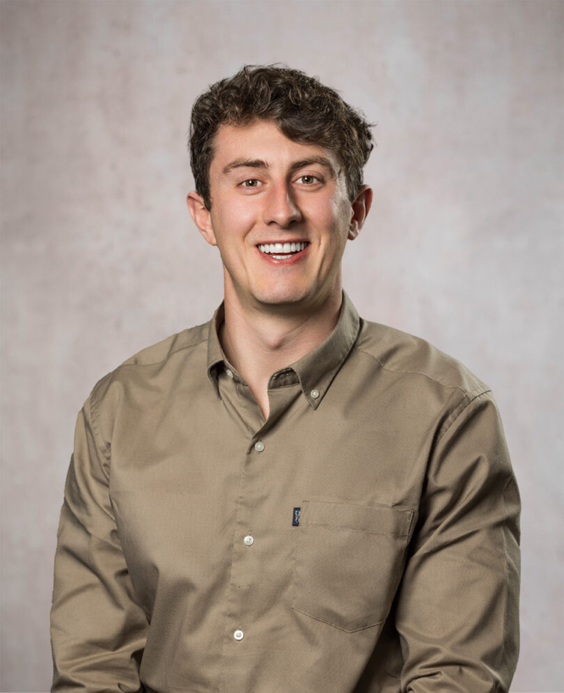 Smiling young man with short curly brown hair wearing a tan button-down shirt against a mottled gray backdrop