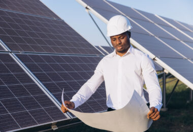 An engineer inspects solar panels at a solar farm while reviewing a blueprint.