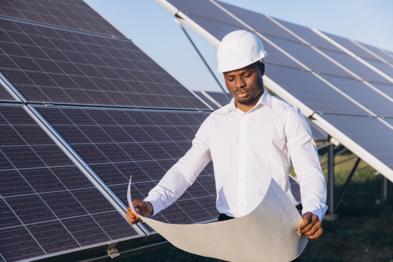 An engineer inspects solar panels at a solar farm while reviewing a blueprint.